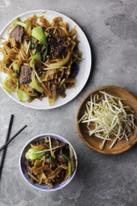 Overview of a bowl of beef chow fun, a plate of beef chow fun and a side of bean sprouts.