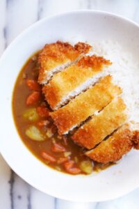 Bird's view of fried chicken cutlet cut into strips served over steamed rice and Japanese curry sauce in a white bowl on marble table.