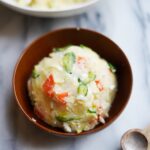 An image showing a bowl filled with Korean potato salad, featuring potatoes, carrots, cucumber, hard-boiled eggs, and diced Fuji apple, all mixed together with creamy mayo dressing.